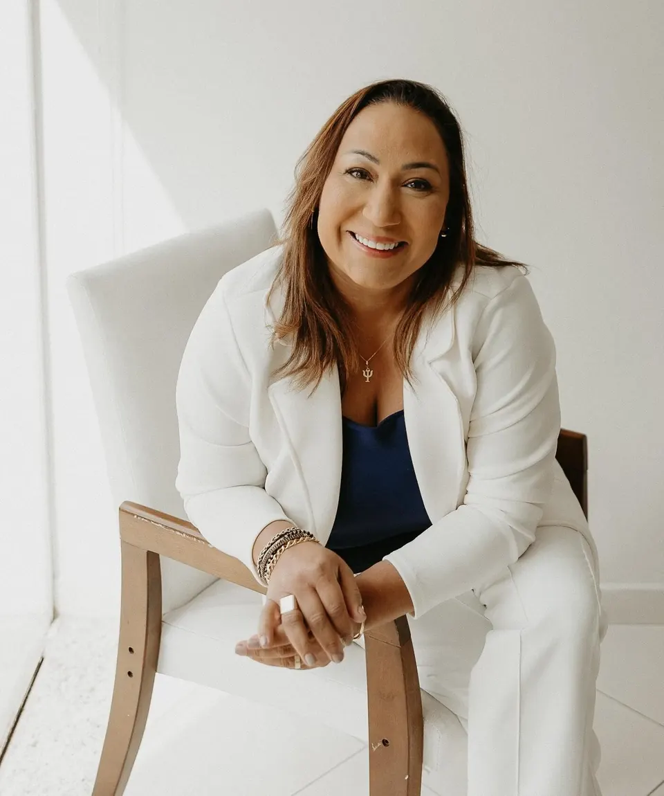 Ana Paula Guimarães sorrindo, de blazer branco, sentada em uma cadeira de madeira clara em um ambiente todo branco.