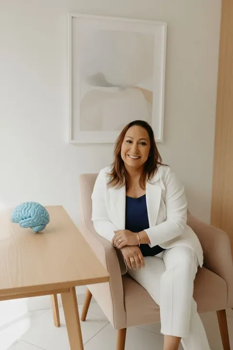 Ana Paula no consultório, sentada à mesa de madeira clara com um modelo de cérebro em cima.