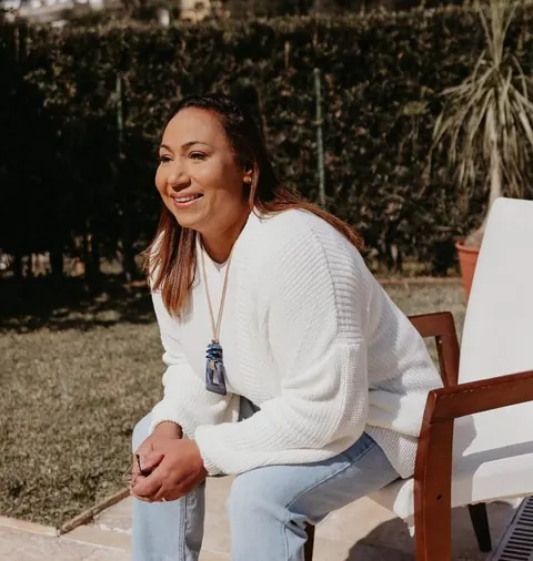 Ana Paula de suéter branco, sentada ao ar livre, sorrindo, em uma cadeira de madeira no jardim.