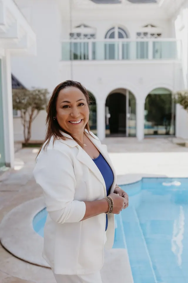 Ana Paula de blazer branco à beira de uma piscina, com uma casa branca de janelas em arco ao fundo.